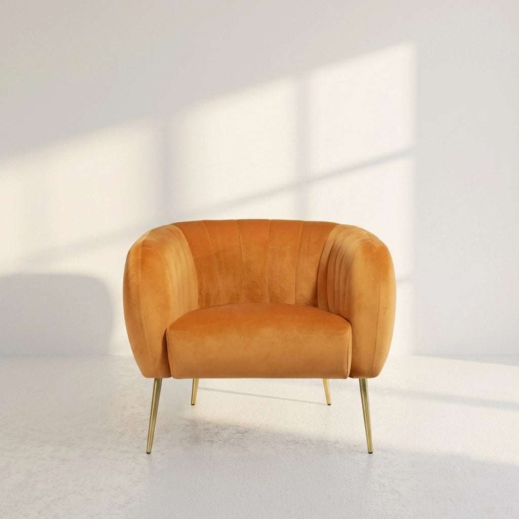 A modern barrel-style armchair upholstered in warm camel velvet with slim gold metal legs, set against a minimalist white wall with soft window-box shadows.