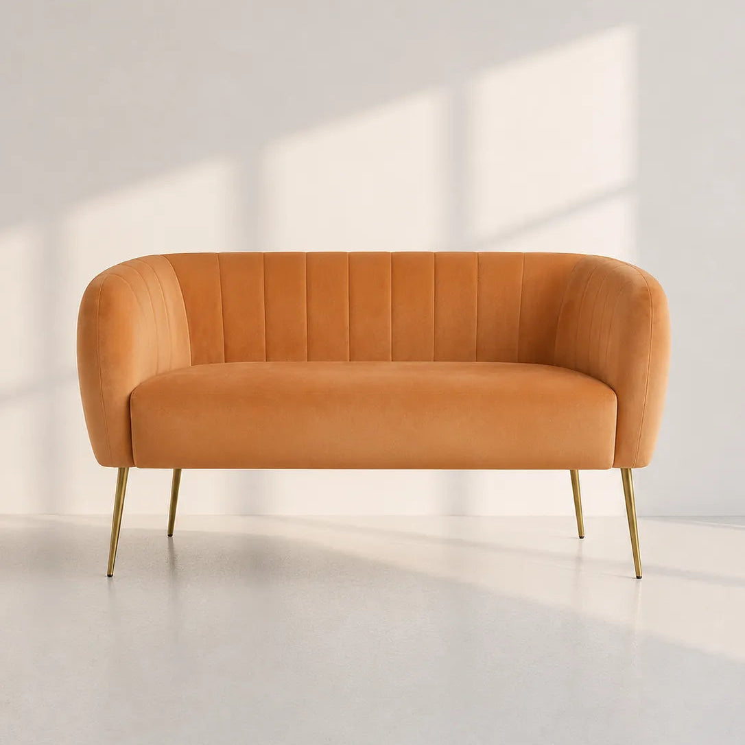 A modern orange velvet loveseat with gold legs, set against a minimalist white wall with soft window shadow patterns.