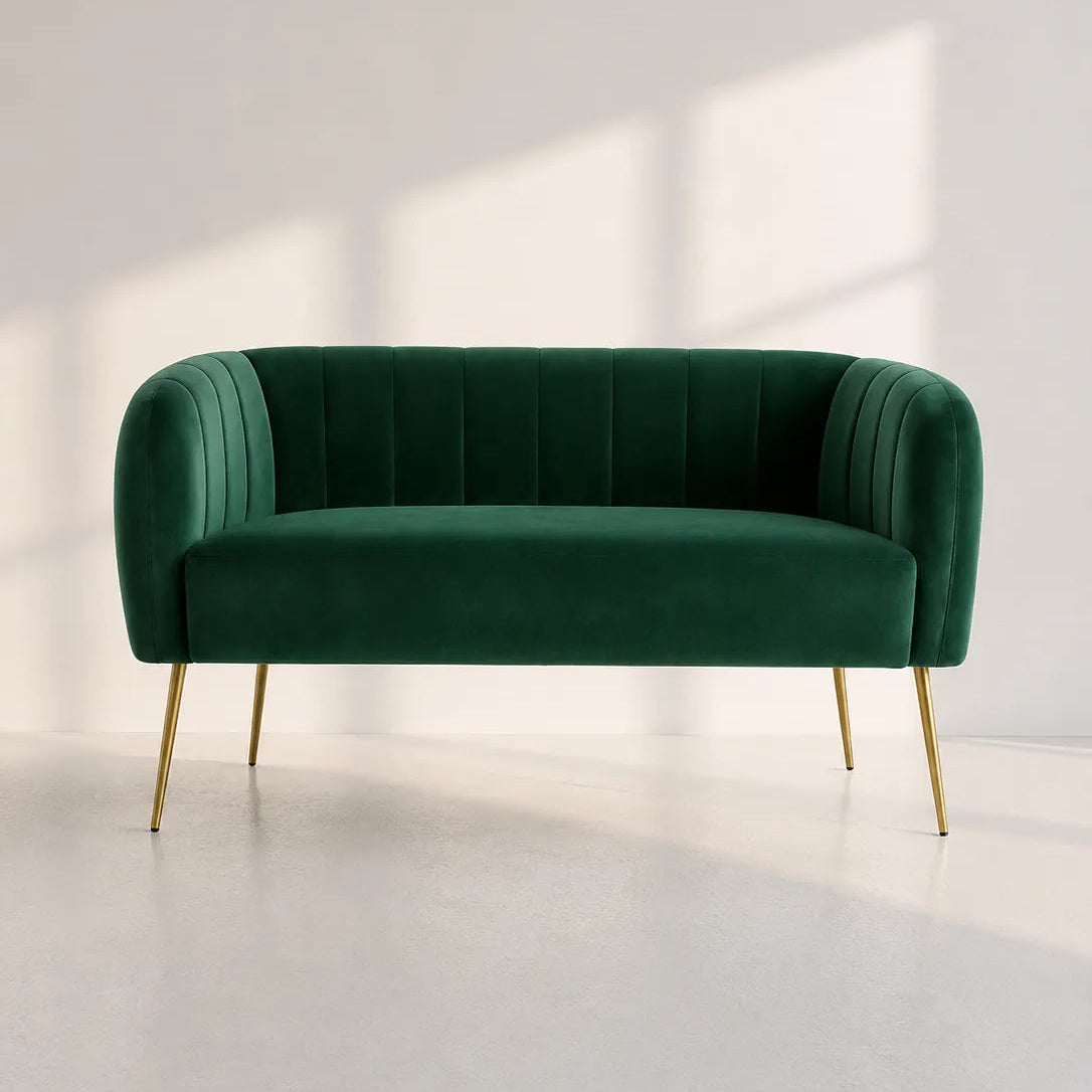 Image of A modern two-seater loveseat sofa upholstered in deep emerald green velvet with vertical channel tufting on the backrest and curved armrests, supported by slim gold-toned metal legs, set against a minimalist off-white wall with soft window-box shadows.