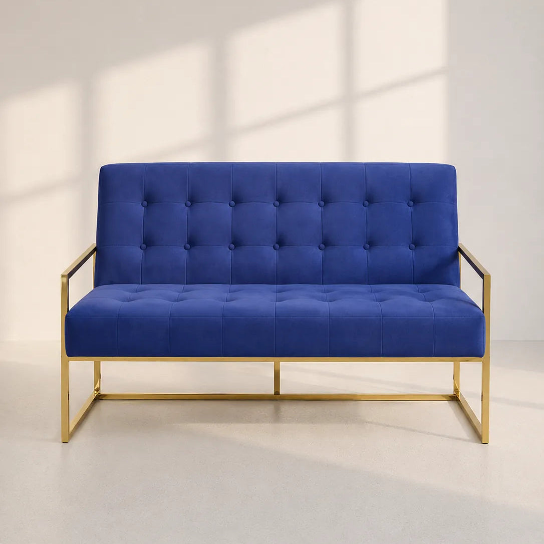 Image of A modern two-seater loveseat sofa with a deep tufted backrest and seat cushion in royal blue velvet, supported by a slim gold-finished metal frame, set against a light gray wall with soft window shadow patterns.