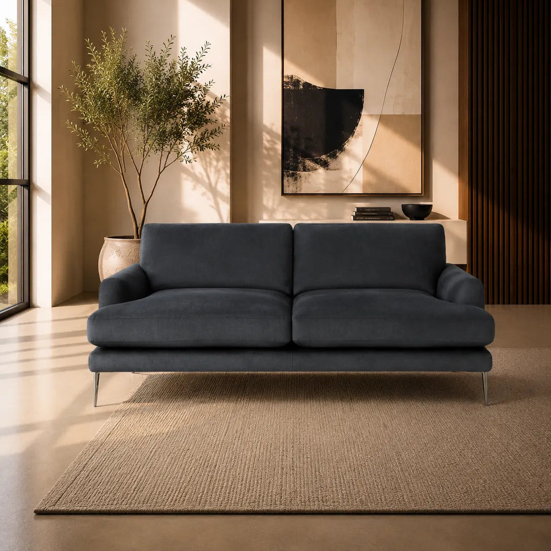 A modern two-seater sofa in dark blue-gray velvet, positioned on a textured beige rug in a contemporary living room with warm ambient lighting, a potted plant, and abstract wall art.