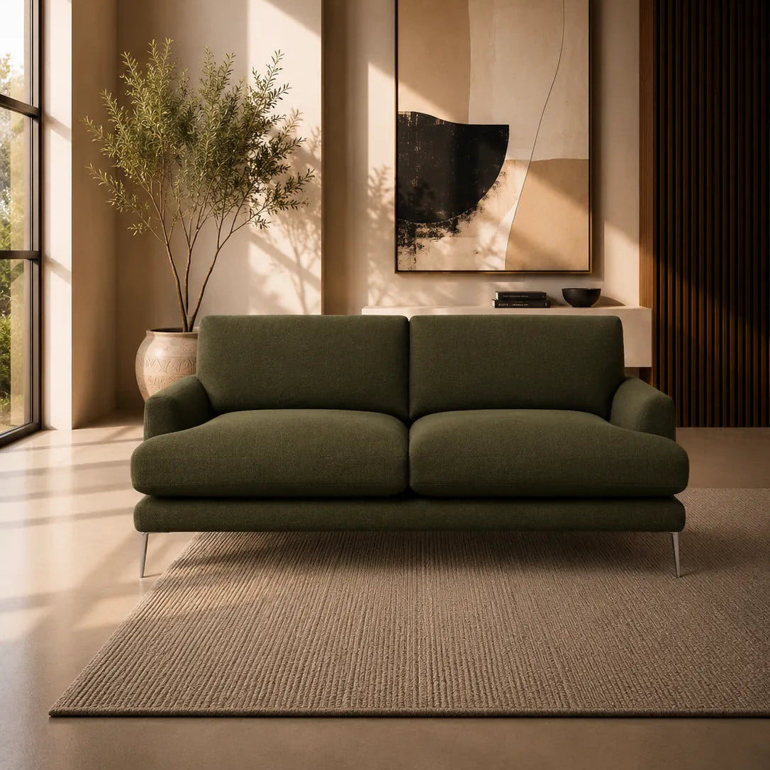 A modern two-seater sofa in olive green fabric, positioned on a textured beige rug in a warm, minimalist living room with a large abstract painting and a potted plant.