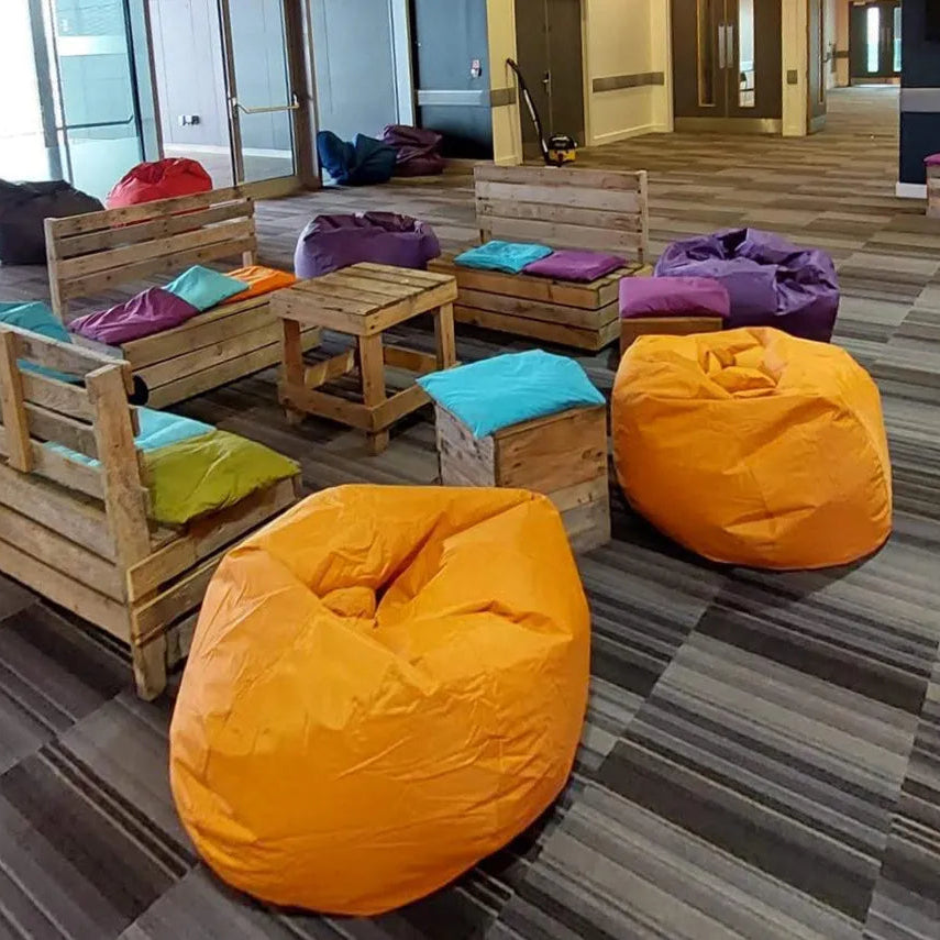 Image of A children's lounge area featuring colorful bean bag chairs in orange, purple, teal, and red, arranged with rustic wooden pallet benches and tables on a gray patterned carpet.