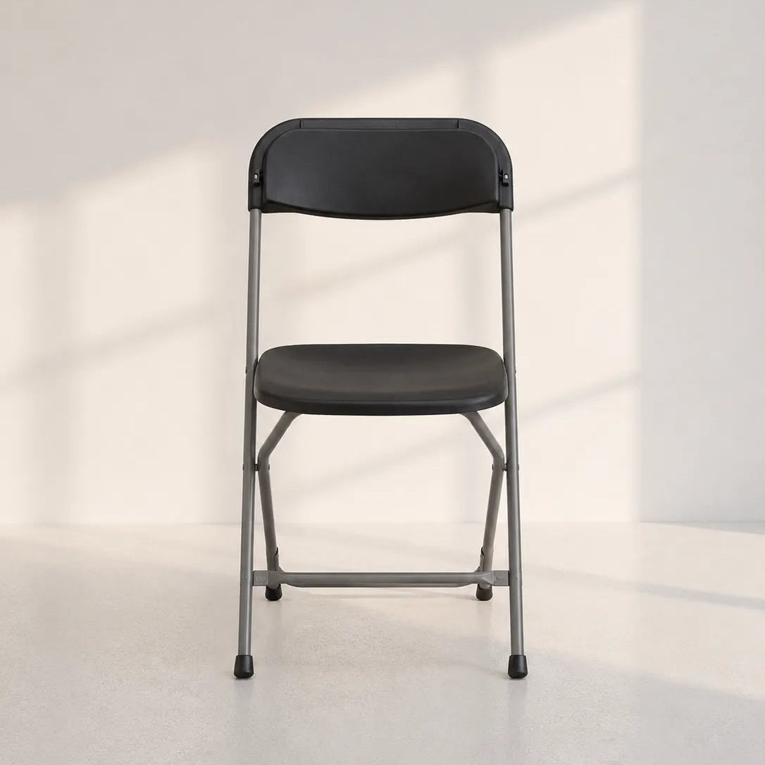 A black folding chair with a gray metal frame, positioned centrally against a minimalist off-white wall with soft window shadow patterns.