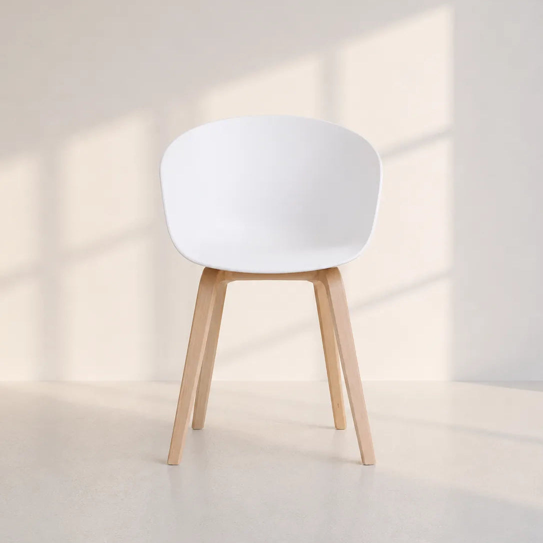 Image of A modern white molded plastic chair with four tapered natural wood legs, set against a minimalist off-white wall and floor with soft window shadow projections.