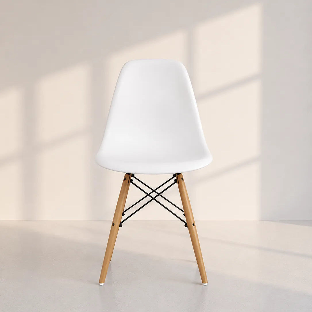 Image of A modern white molded plastic side chair with light natural wood legs and black wire cross-bracing, set against a minimalist beige wall with soft window shadow patterns.