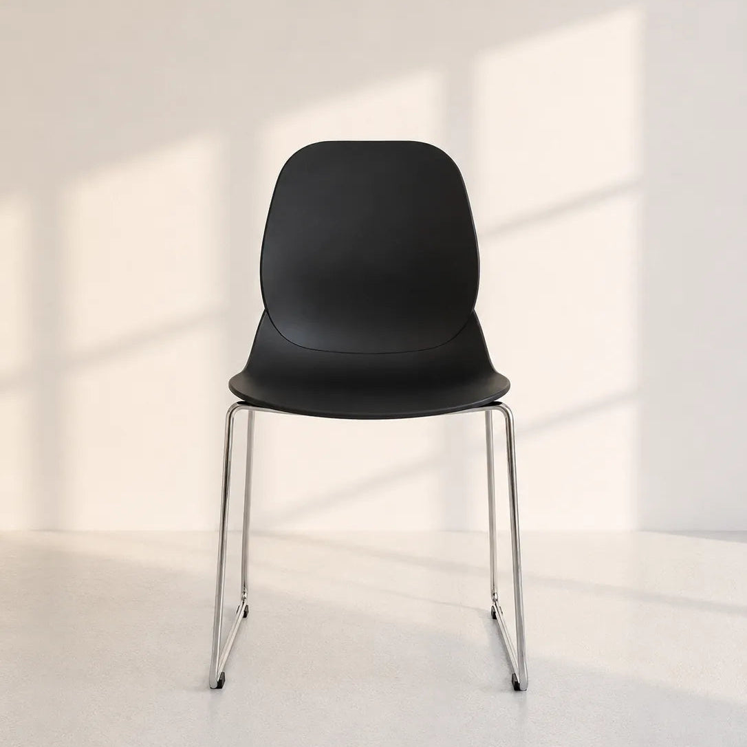 A modern black molded plastic chair with slim chrome legs, positioned in a minimalist white room with soft window shadow patterns on the wall.