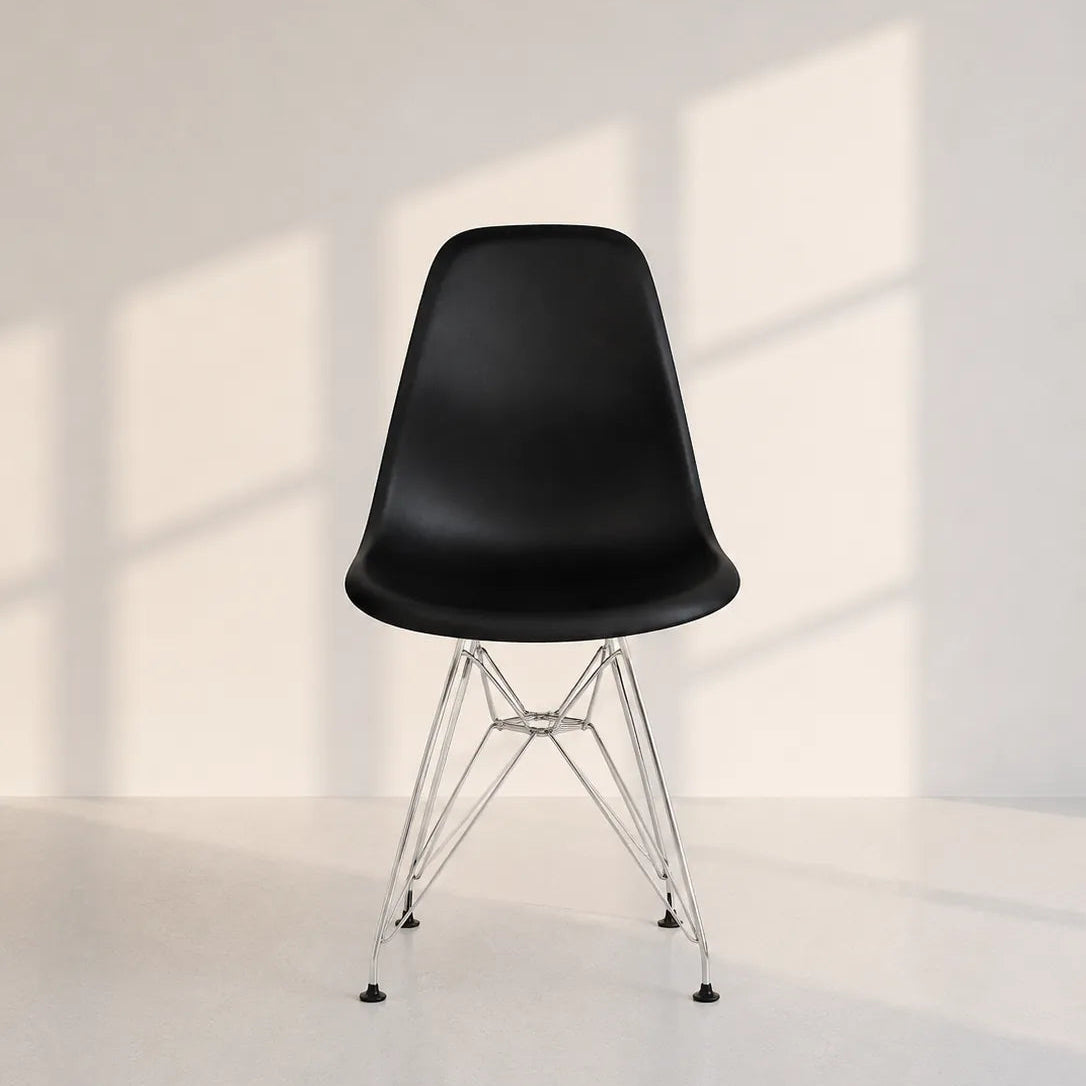 Image of A modern black molded plastic side chair with a chrome wire pedestal base, set against a minimalist off-white wall with soft window shadow patterns.