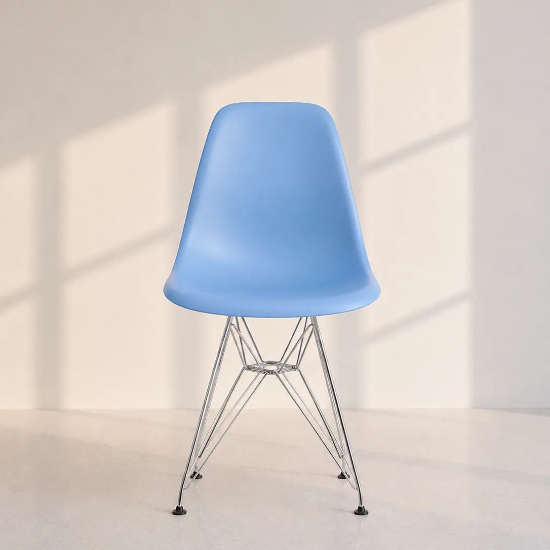 A light blue molded plastic side chair with a chrome wire four-leg base, set against a warm off-white wall with soft window shadow patterns.