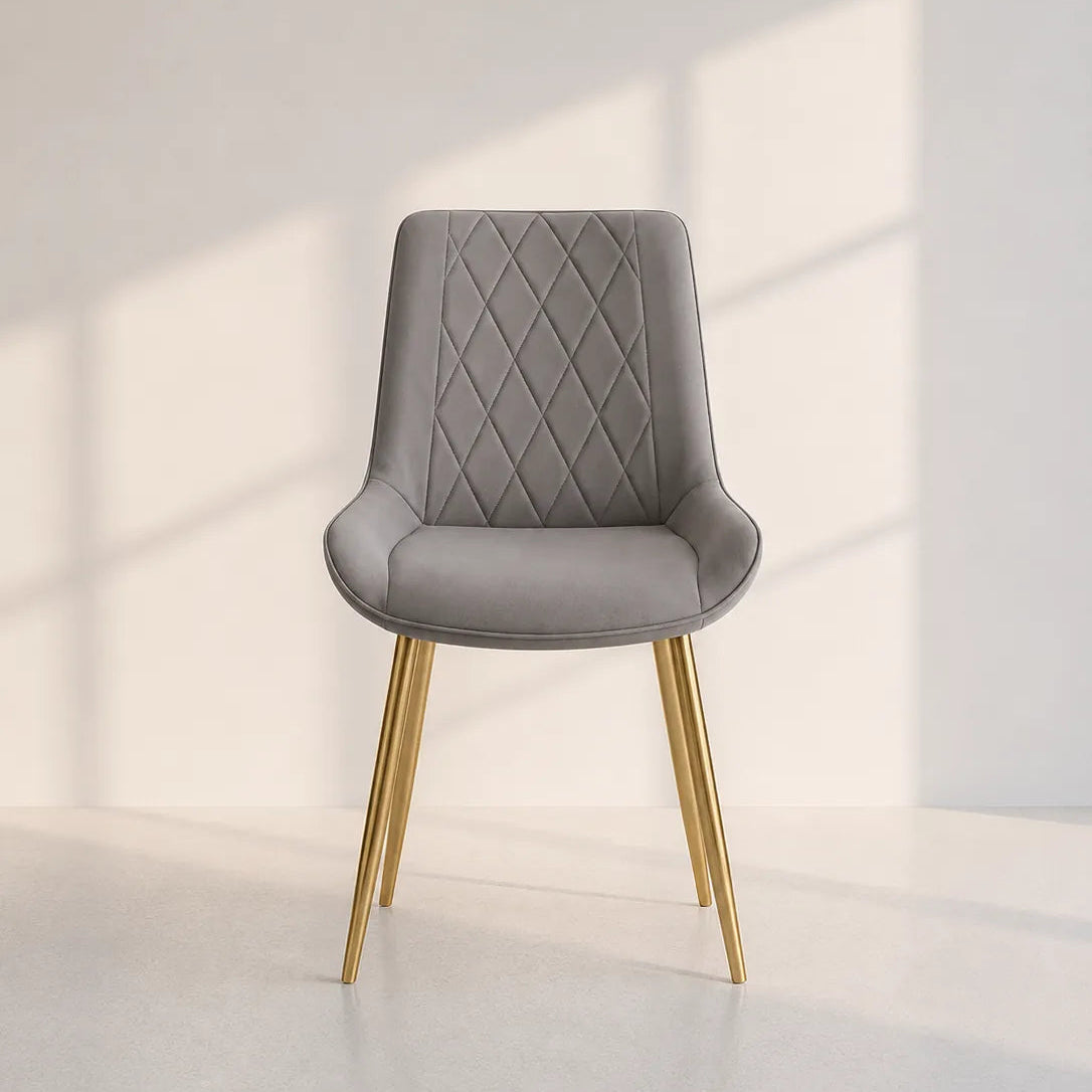 A modern gray upholstered dining chair with a diamond-quilted backrest and tapered gold metal legs, set against a minimalist off-white wall with soft window shadow projections.