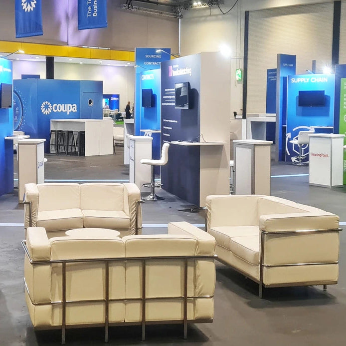 Image of A set of modern cream-colored leather or faux-leather lounge chairs and sofas arranged in a large exhibition hall. The furniture features clean lines, low-profile backs, and slim metallic frames.