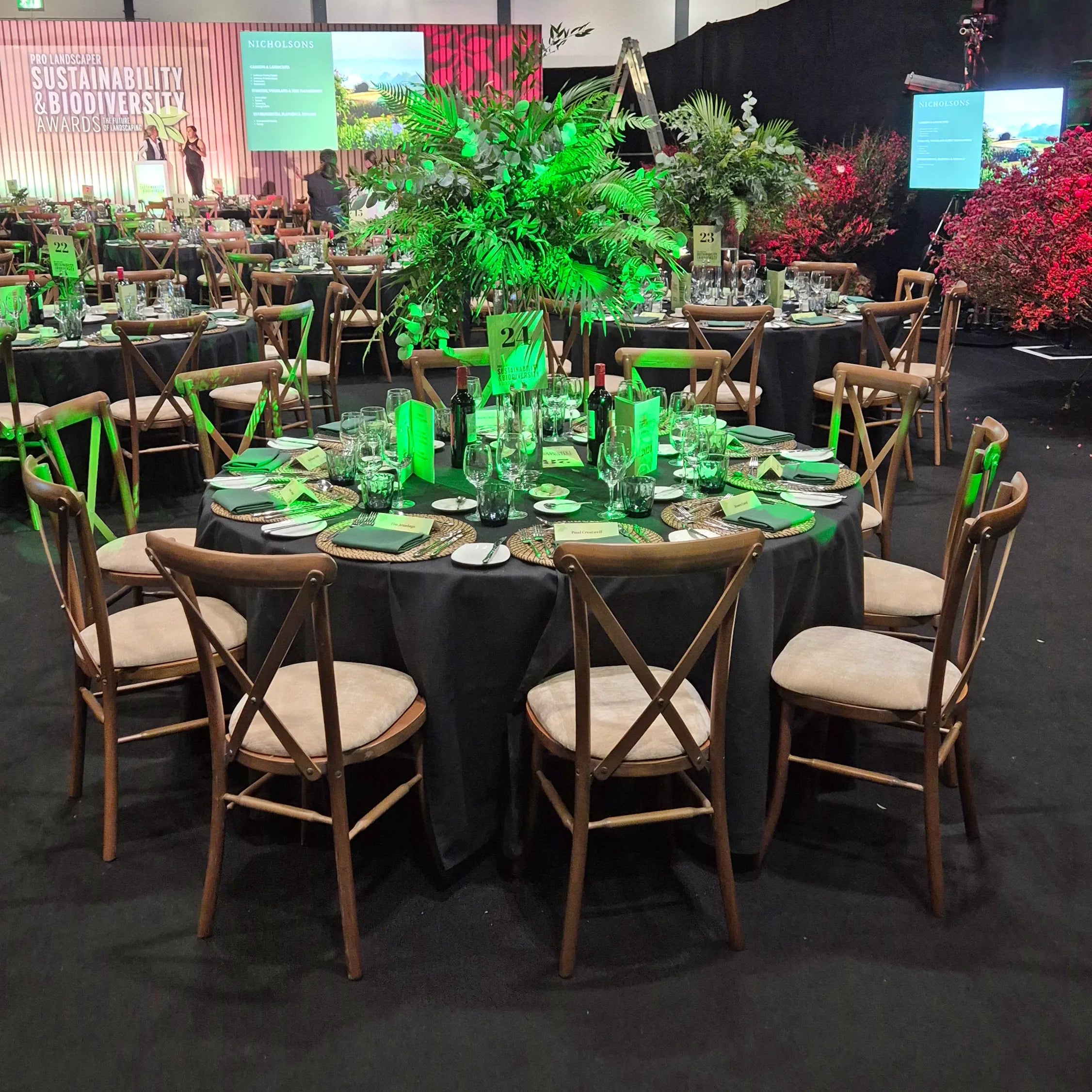 Image of A large event space setup with multiple round tables covered in black tablecloths, surrounded by wooden chairs with beige seat cushions. The tables are set with green napkins, plates, and glassware, and decorated with tall green foliage centerpieces.