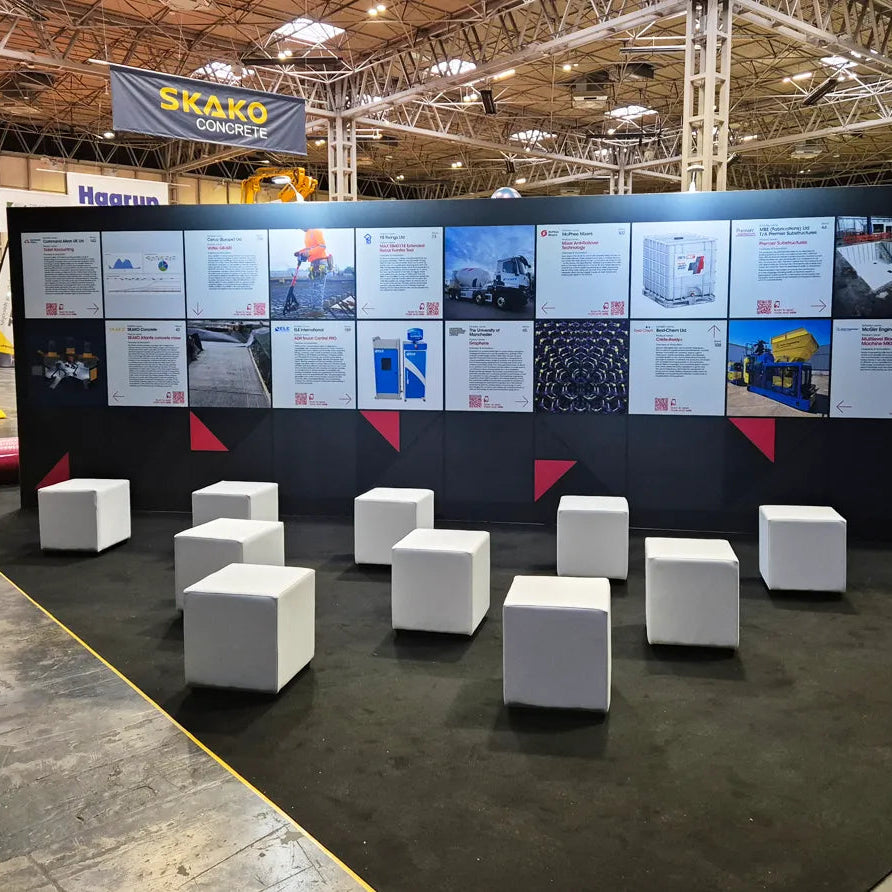 A large exhibition-style display wall with multiple printed panels and several white cube stools arranged in front on a dark floor inside a convention hall.