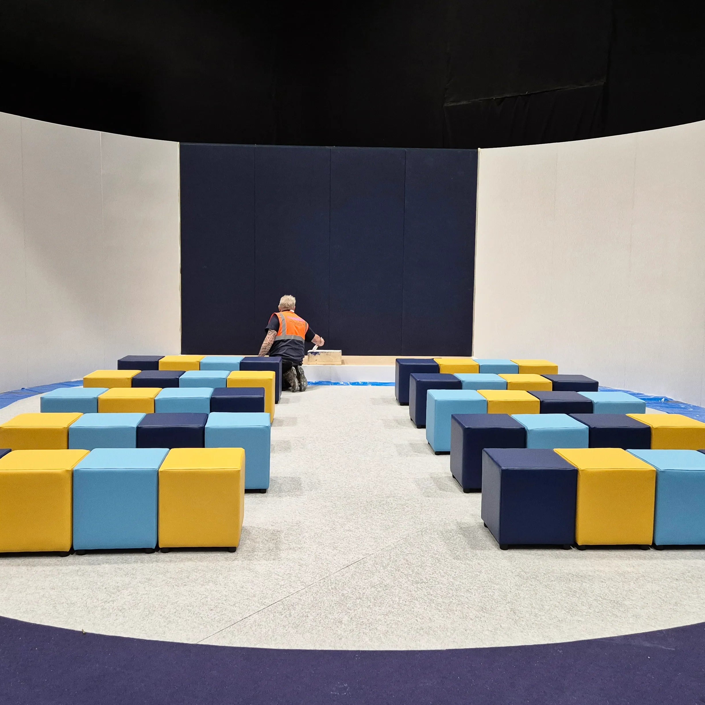 Image of A large indoor play area or gymnasium with a circular floor setup, featuring rows of colorful modular foam blocks in navy, light blue, and yellow. A person in an orange shirt is seated near the center, with a dark navy back wall and white side walls behind them.