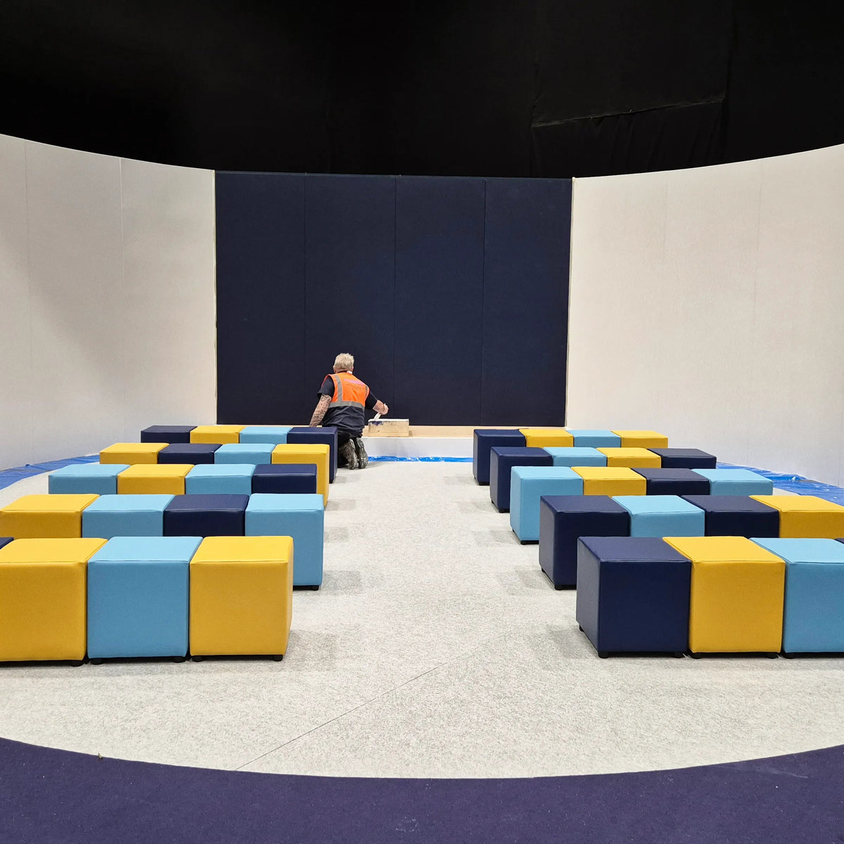 A large indoor play area or gymnasium with a circular floor setup, featuring rows of colorful modular foam blocks in navy, light blue, and yellow. A person in an orange shirt is seated near the center, with a dark navy back wall and white side walls behind them.