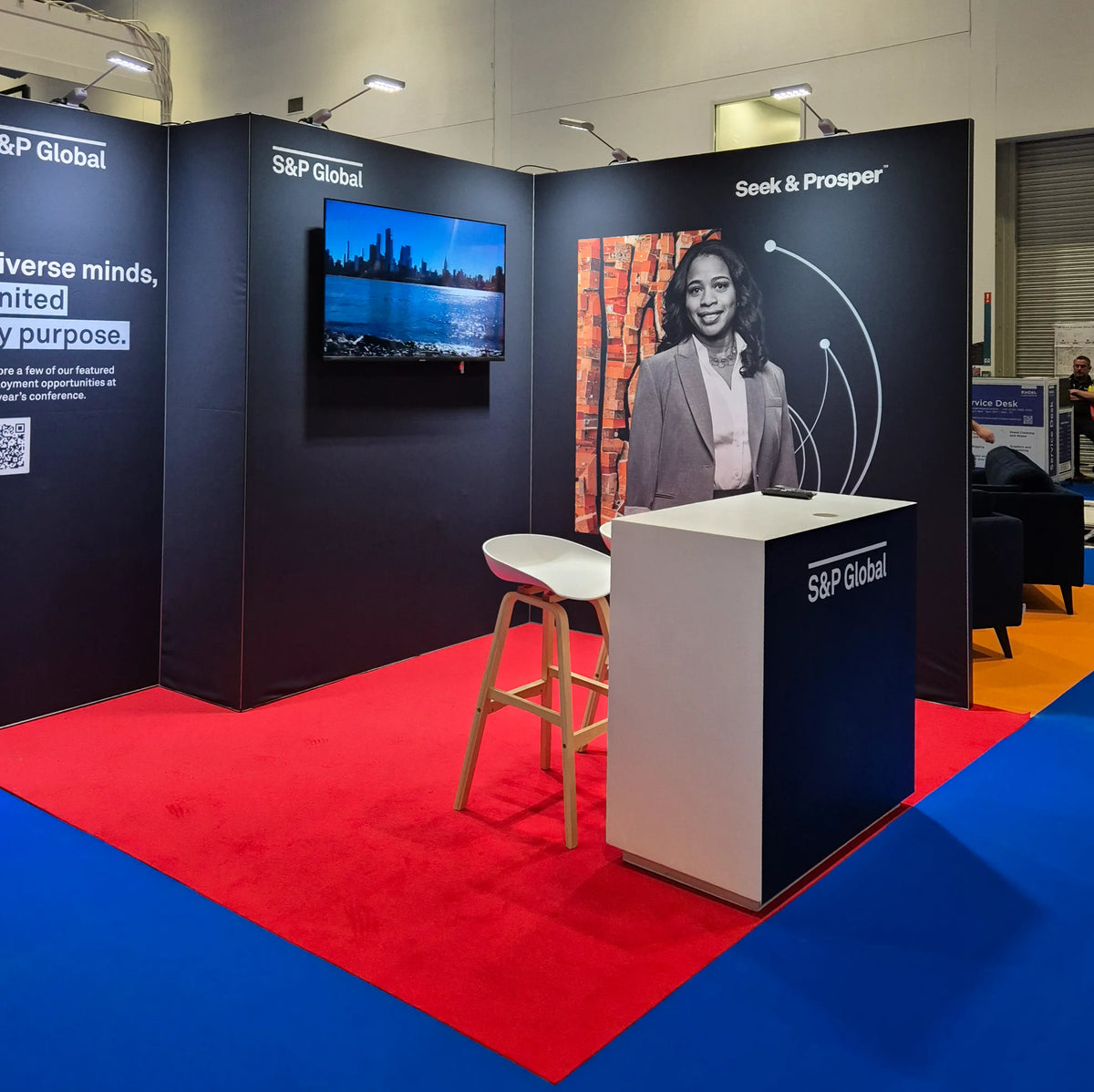 S&P Global exhibition booth at a trade show, featuring a white and navy counter, a white stool, a wall-mounted screen, and branded display panels on a red and blue floor.