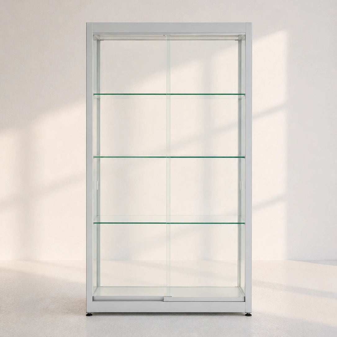 A tall, narrow glass display cabinet with a white frame and four horizontal glass shelves, set against a minimalist white wall with soft window shadows.