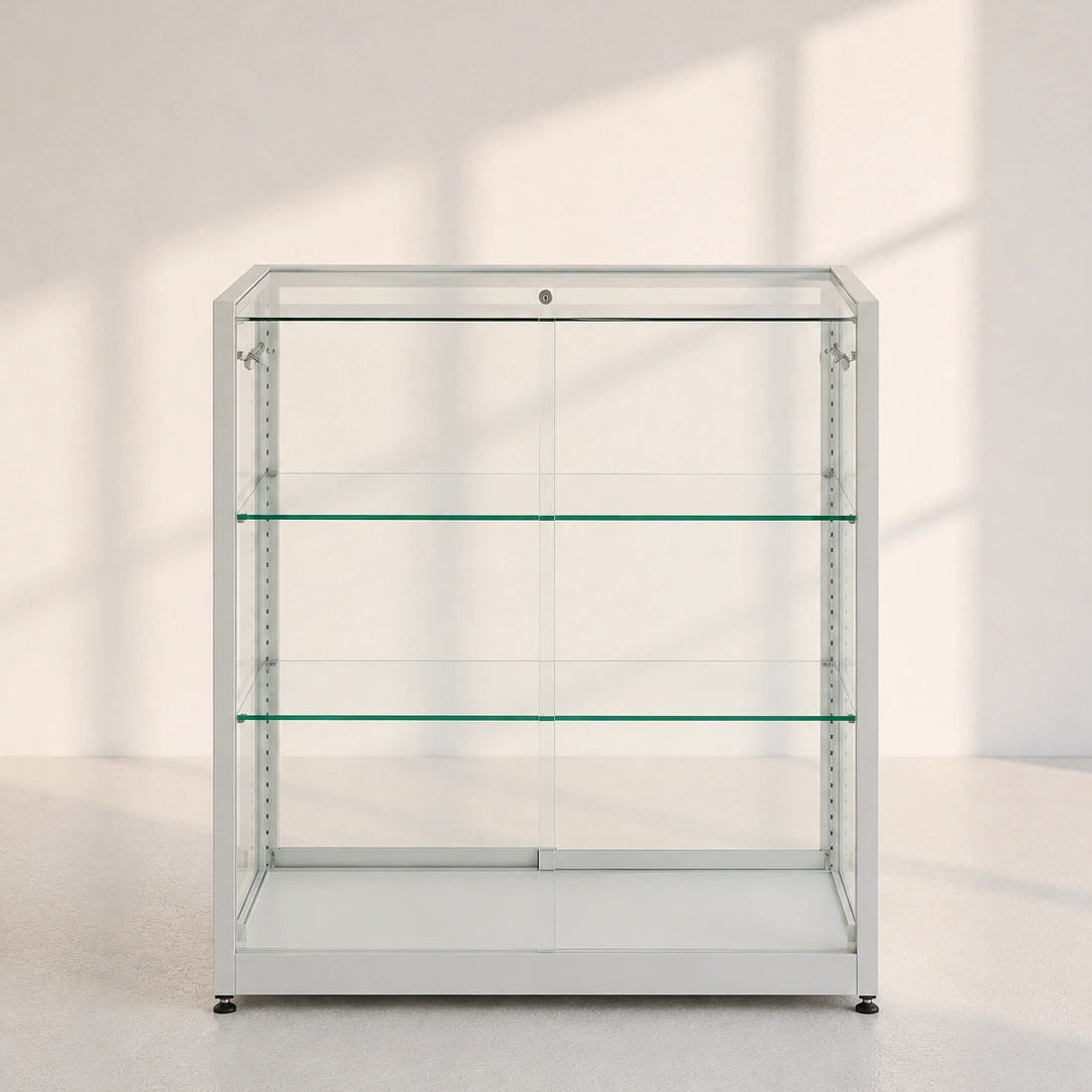 Image of A tall, empty glass display case with three shelves and a silver metal frame, set against a minimalist off-white wall with soft window shadow patterns.