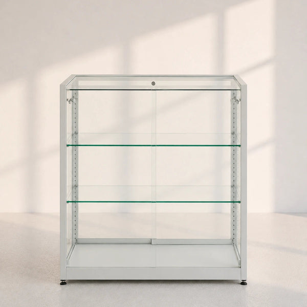 A tall, empty glass display case with three shelves and a silver metal frame, set against a minimalist off-white wall with soft window shadow patterns.