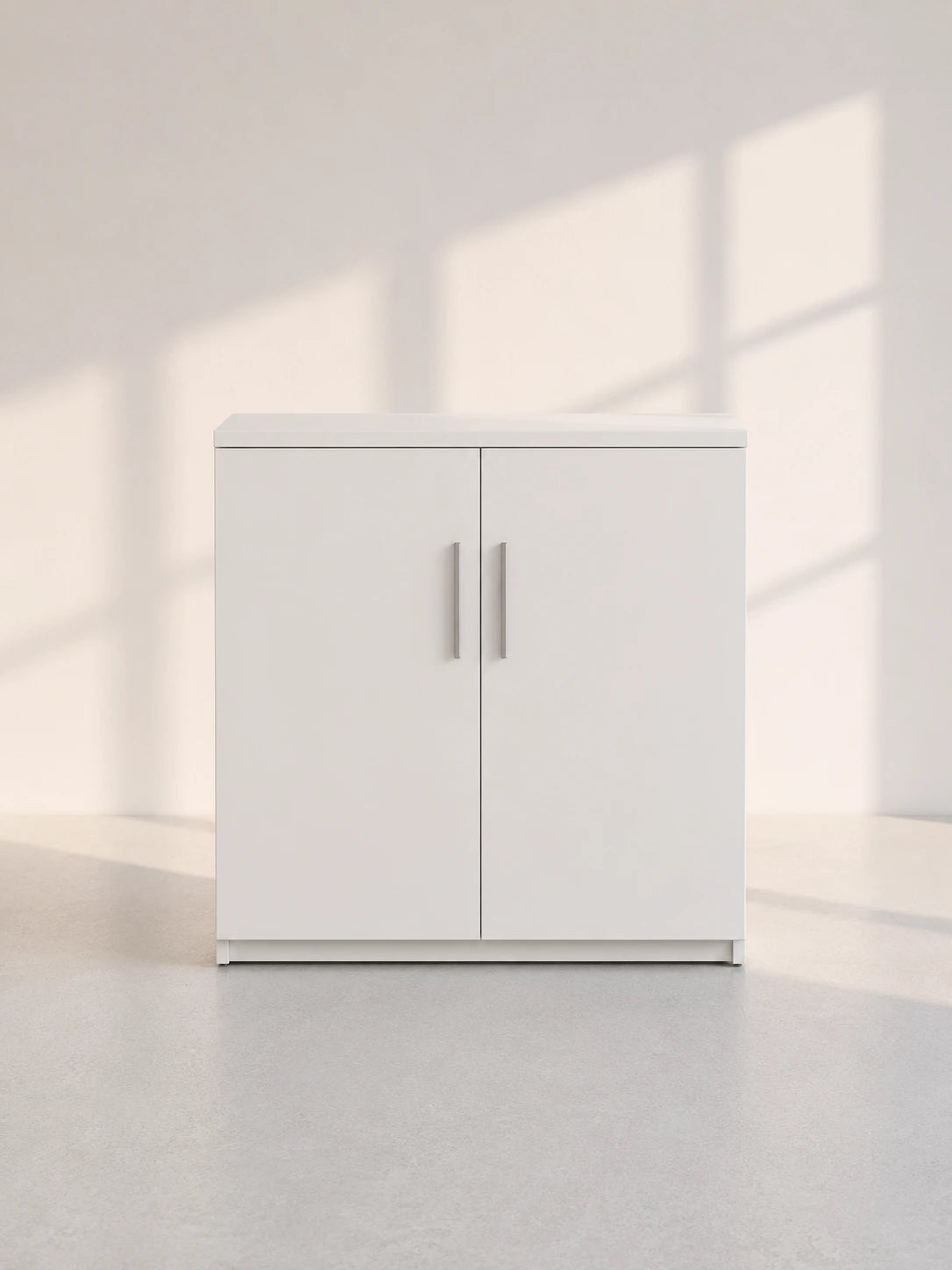 Image of A modern white two-door cabinet with slim silver handles, set against a minimalist off-white wall with soft window shadow patterns and a light gray floor.