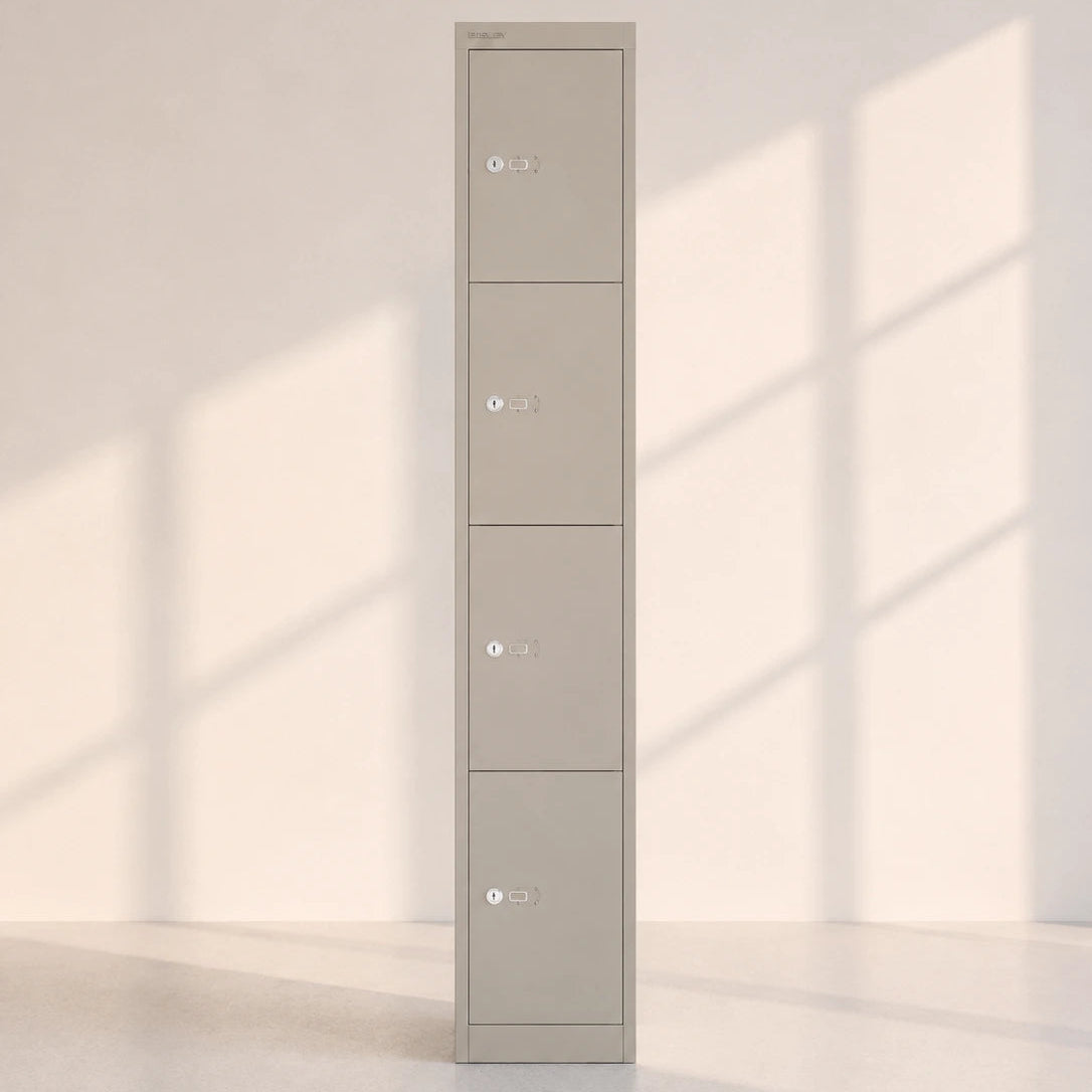 Image of A tall, narrow four-door metal locker in a warm gray or taupe color, standing against a minimalist off-white wall with soft window shadow patterns cast across it.