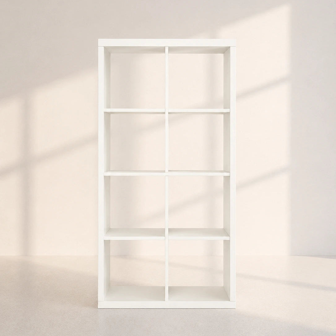 A tall white open shelving unit with six square compartments, photographed against a warm off-white wall with soft window-shadow light.