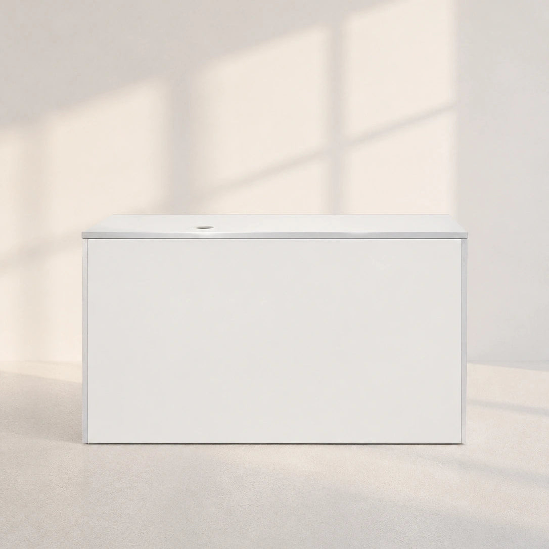 A minimalist white rectangular box with a small circular detail on top, set against a warm beige wall and floor with soft window shadow projections.