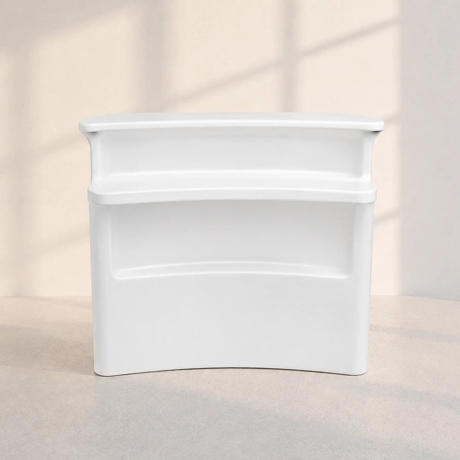 A white plastic two-tier storage organizer with a curved bottom edge, set against a warm beige wall with soft window shadow patterns.