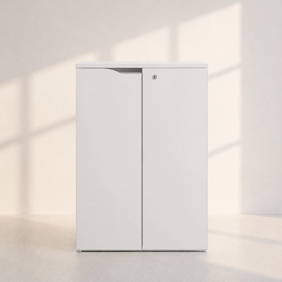 A tall, minimalist white two-door storage cabinet with a small round lock, set against a light beige wall with soft window shadow patterns.