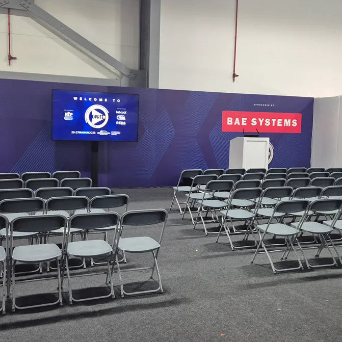 Image of An indoor event space with rows of black folding chairs facing a blue stage backdrop, a large screen, and a red BAE SYSTEMS sign.