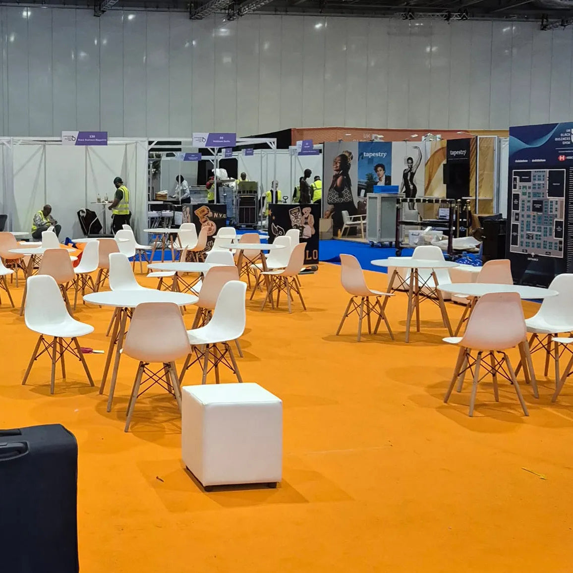 Image of A large exhibition hall with many white dining tables and chairs arranged on a bright orange carpet, with a few people and booths visible in the background.