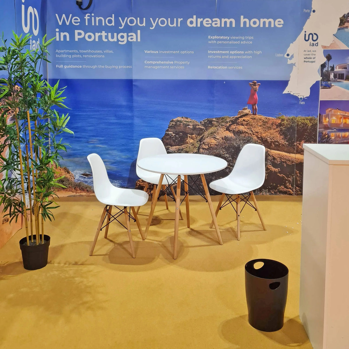A modern 3-piece dining set consisting of a round white table and two white chairs with light wood legs, displayed on a yellow carpet in front of a large blue promotional banner for real estate in Portugal.