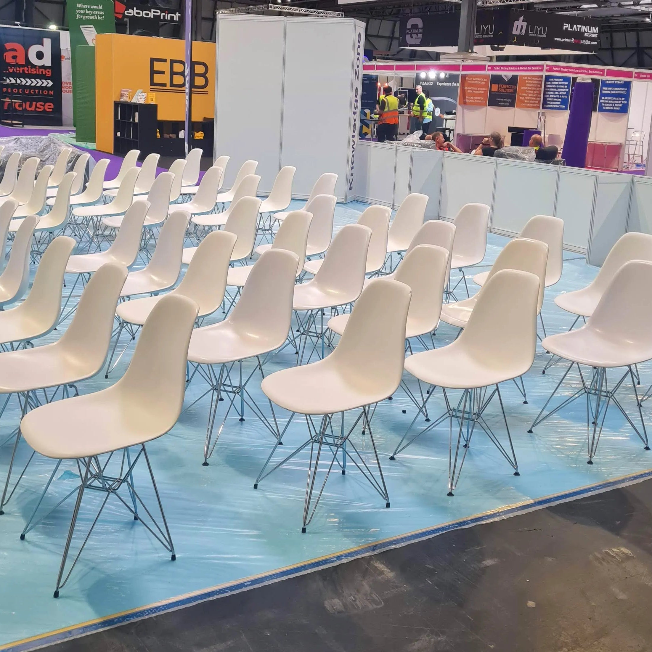 Image of Multiple white molded plastic chairs with metal wire legs arranged in rows on a light blue protective floor covering at a trade show or exhibition.
