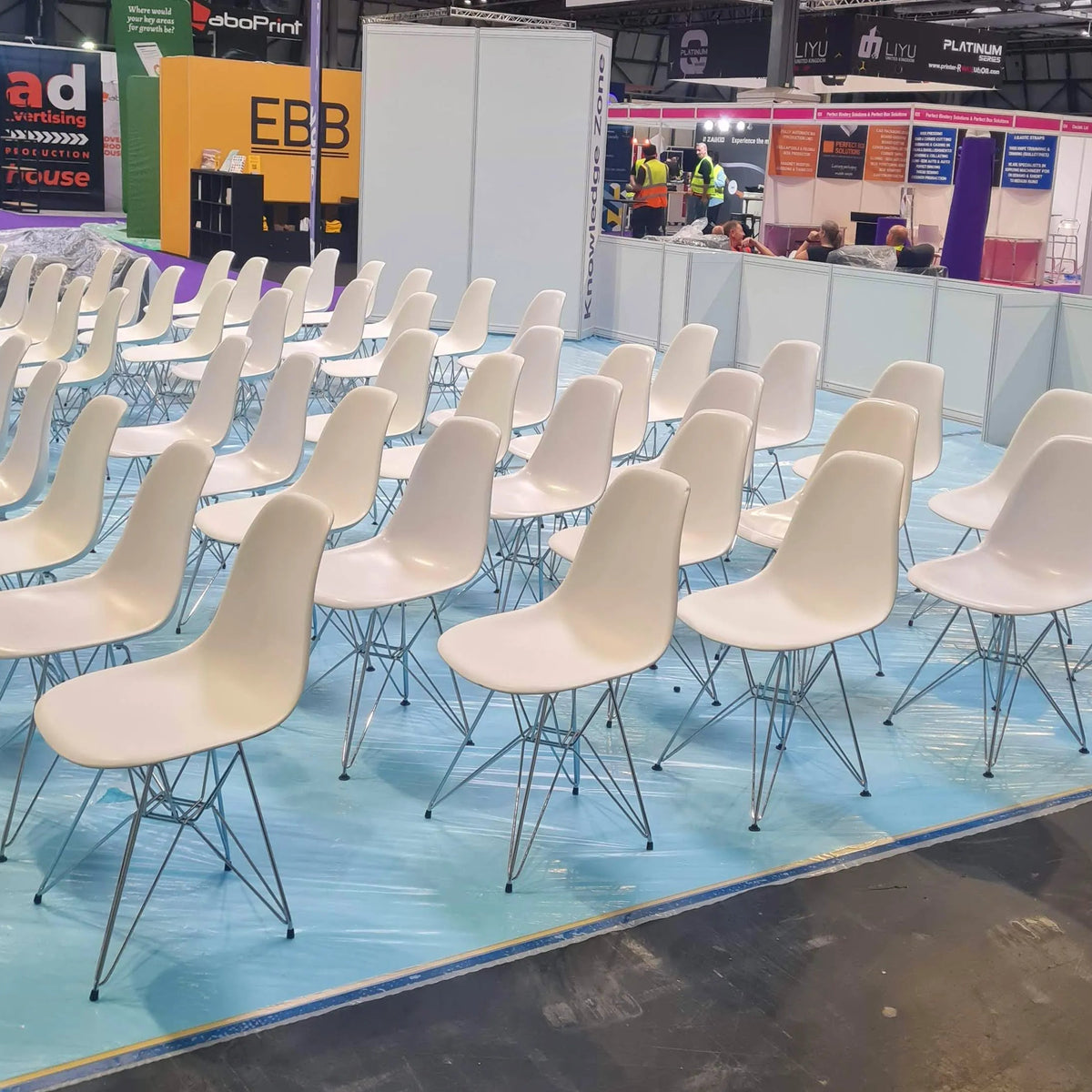 Multiple white molded plastic chairs with metal wire legs arranged in rows on a light blue protective floor covering at a trade show or exhibition.