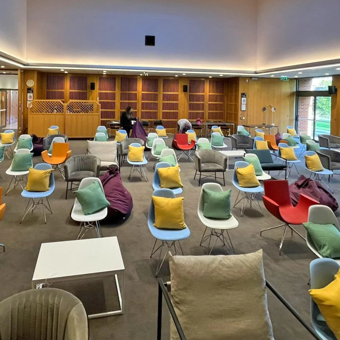 Image of A large modern indoor meeting or lounge space with rows of colorful chairs, some with yellow and green pillows, arranged in a spacious room with wood paneling and windows.