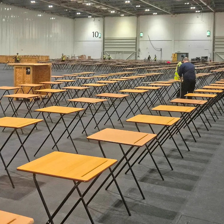 Image of Rows of portable wooden desks with orange-tan tops and black metal legs arranged in a large indoor event space or warehouse. A person in a high-visibility vest stands near the right side in the background.