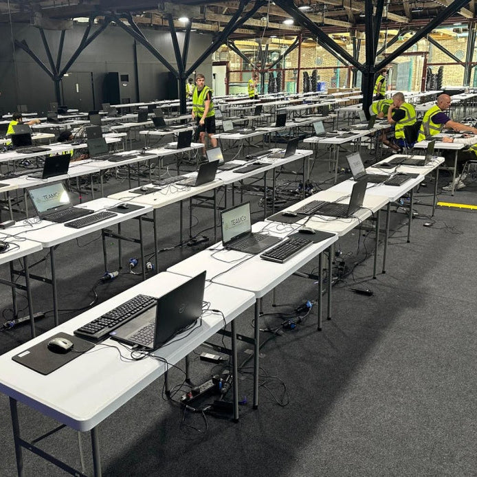 A large indoor warehouse or event space with rows of white folding tables set up with laptops, keyboards, and mice. Several people in high-visibility vests are standing and working around the tables.