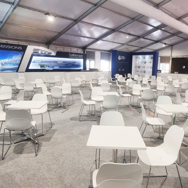 Image of A large event hall or exhibition space with rows of white tables and white chairs arranged under a high industrial-style tent ceiling. Multiple display screens are mounted on the back wall, and branded booths or panels are visible in the background.