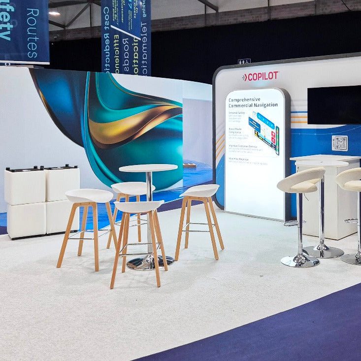 A booth display at an exhibition featuring a white and blue interactive navigation screen labeled 'XCOPILOT', surrounded by white bar stools with light wood legs and small round tables on a curved white platform.