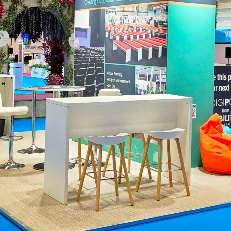 Image of A white bar-height counter with three white bar stools featuring light wood legs, displayed on a light wood floor with a blue carpet border at a trade show booth.