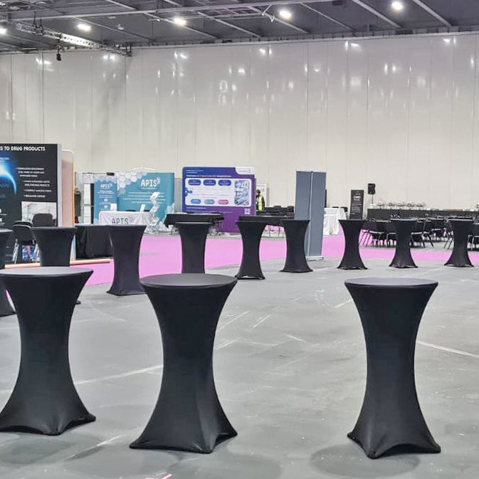 Image of A large exhibition hall with multiple black-covered high tables arranged in rows on a gray floor, with booths and displays visible in the background.