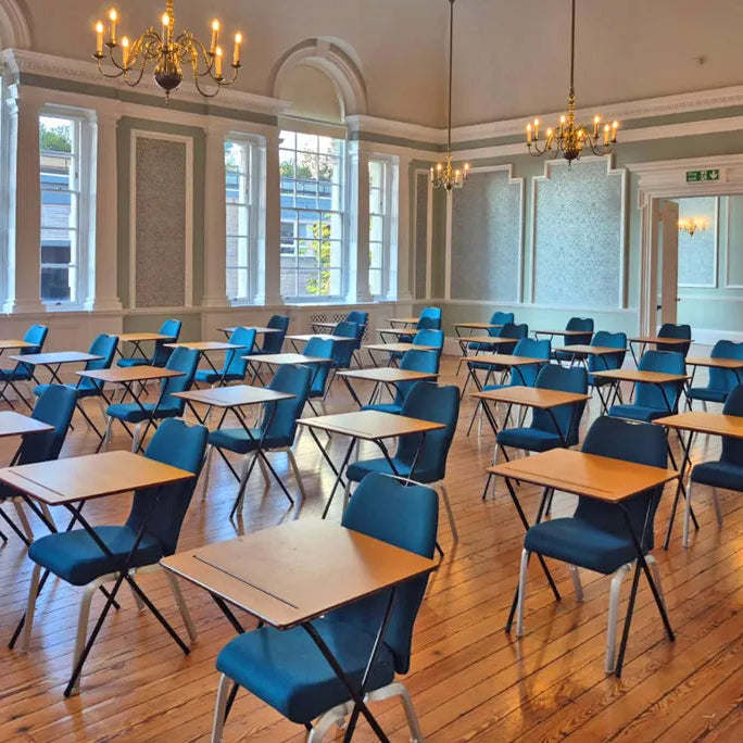 A large classroom or conference room with rows of wooden desks and blue folding chairs arranged on a polished wood floor, with tall windows, chandeliers, and pale green walls.