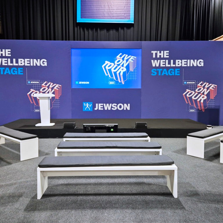 A large event stage setup with a blue branded backdrop reading 'THE WELLBEING STAGE' and 'JEWSON' in the center, plus multiple white benches with dark seat cushions arranged in front on a carpeted floor.