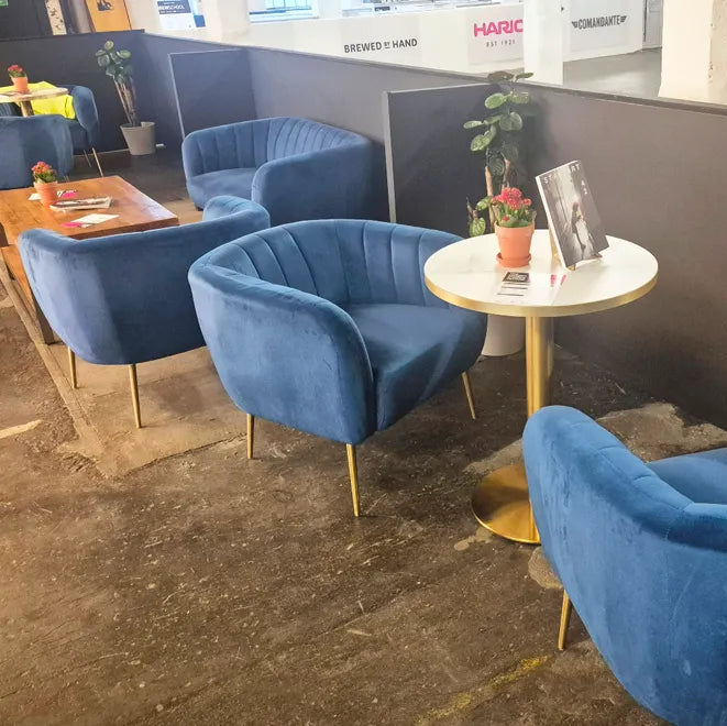 A set of modern blue velvet armchairs with gold legs arranged around a small round white table with a gold base in an office lounge setting.