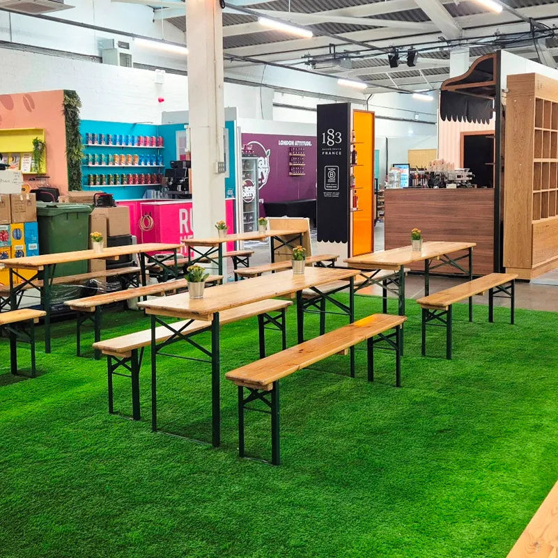 Image of A large indoor event space or cafe area featuring multiple wooden picnic-style tables with matching benches arranged on bright green artificial turf. The background shows a modern interior with shelving, vending machines, and a counter.