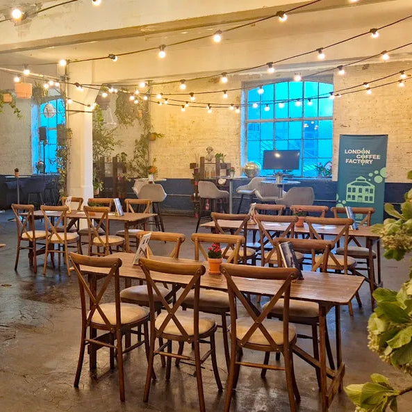A large indoor dining or event space with multiple wooden tables and matching wooden chairs arranged throughout. The room has exposed brick walls, hanging string lights, large windows with blue-tinted glass, and decorative greenery.