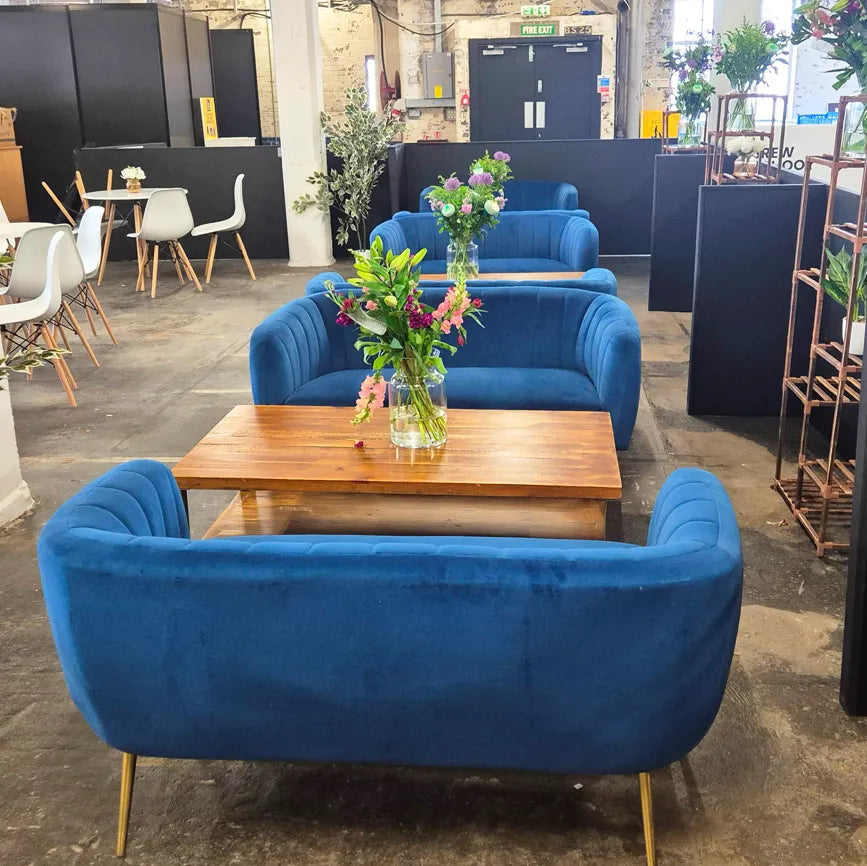 Image of A set of modern blue velvet armchairs with gold metal legs arranged around a wooden coffee table in a loft-style office or cafe setting.