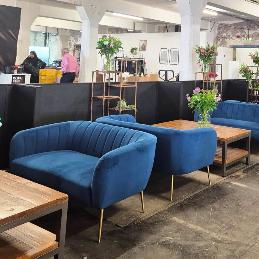 A row of modern blue velvet armchairs and matching sofas arranged with wooden tables in a warehouse-style lounge or retail interior. The furniture has gold-toned metal legs and is styled with floral arrangements.