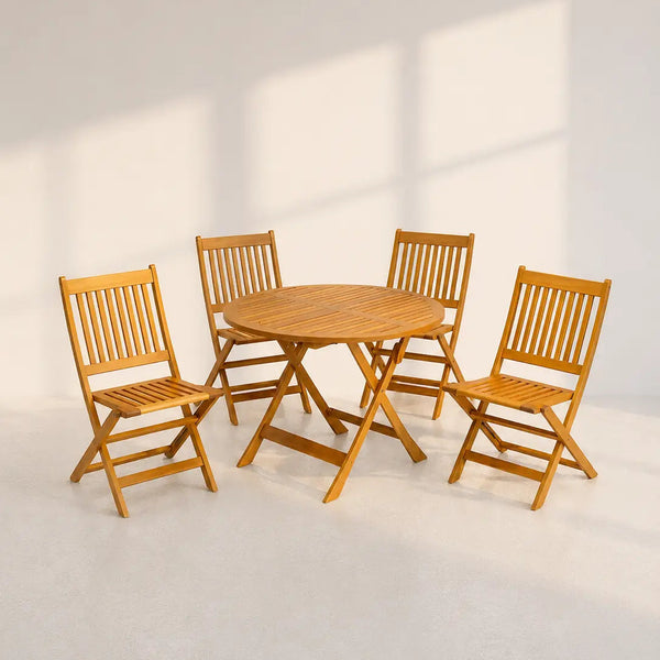 A 5-piece outdoor dining set made of light brown teak wood, including a round folding table and four matching folding chairs, set against a minimalist off-white wall with soft window shadows.