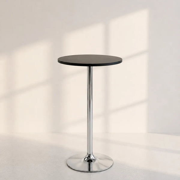 A modern bar-height side table with a round black top and a polished chrome pedestal base, set against a minimalist off-white wall with soft window shadow patterns.